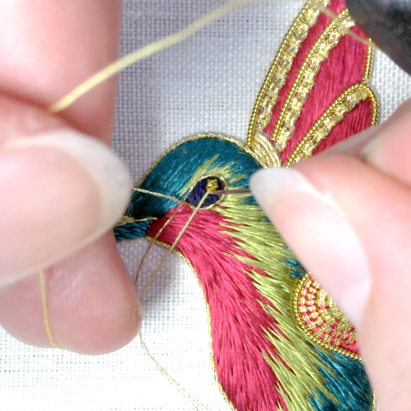 hands stitching a chip of bright check onto the eye of an embroidered hummingbird